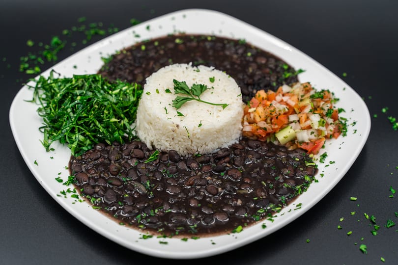 A white plate with black beans, white rice, green collard greens, and pico de gallo.