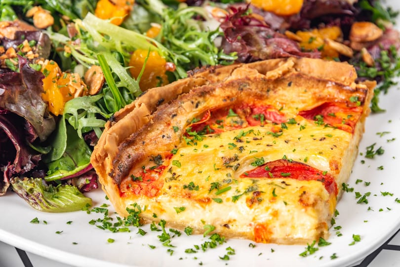 Slice of quiche with tomatoes and herbs, served with a fresh green salad with oranges.
